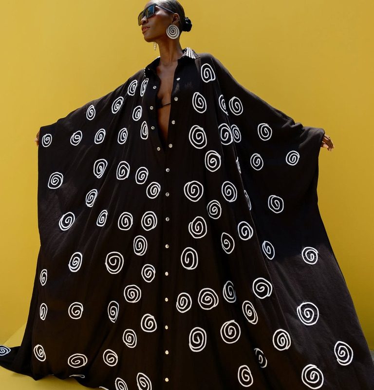 Female model in Cute Saint black oversized maxi shirt dress with white spiral adire print, arms outstretched against yellow background, Irin-Ajo Resort 2025