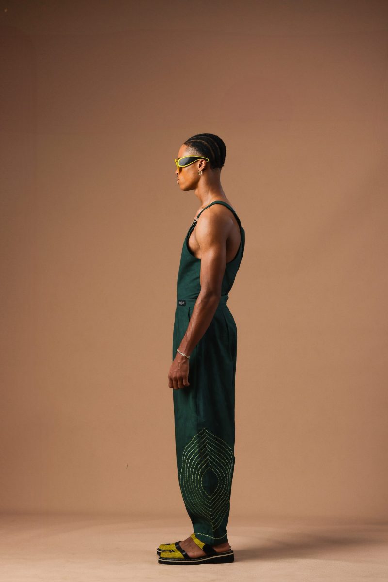 Side view of a model wearing the Cute-Saint Riri Dungaree in dark forest green, featuring a sleeveless top and wide-leg trousers with yellow contrast stitching on the lower legs.