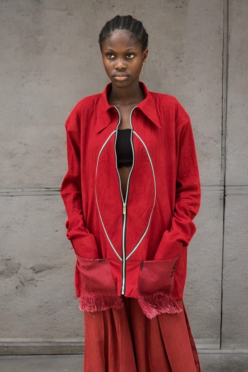 Female model in Cute Saint Ewa red linen zip jacket with oval piping detail and fringe-trimmed patch pockets, open over black bralette, paired with rust red ultra wide-leg trousers