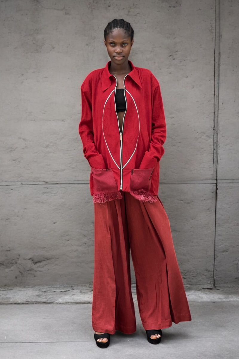 Female model in Cute Saint Ewa red linen zip jacket with oval piping detail and fringe-trimmed patch pockets, open over black bralette, paired with rust red ultra wide-leg trousers
