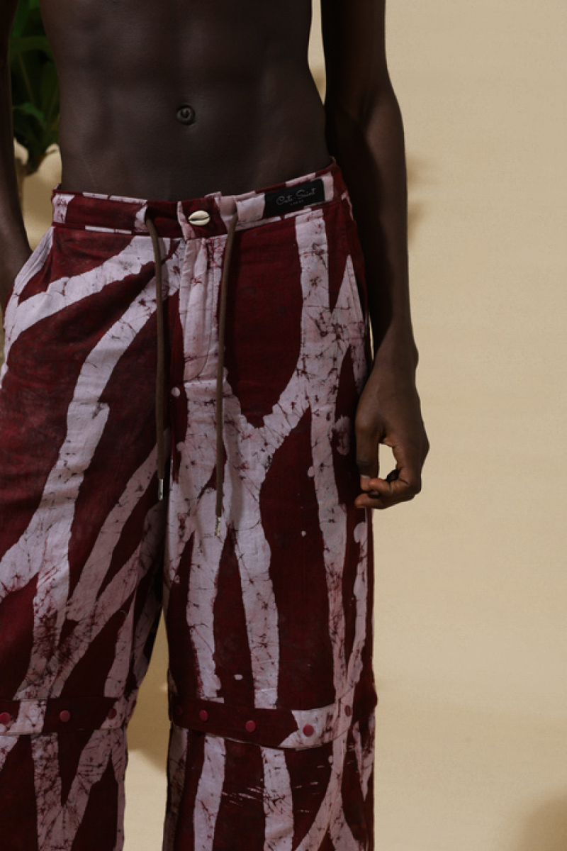 Close-up of shirtless male model wearing Cute Saint Irin-Ajo burgundy and white stripe batik wide-leg trousers showing Cute Saint branded waistband, cowrie shell button, and drawstring detail