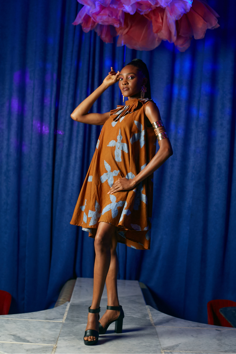 Female model on runway wearing Cute Saint rust brown sleeveless swing mini dress with oversized blue bird batik print and ruffled neckline, styled with gold arm cuffs and black heeled sandals