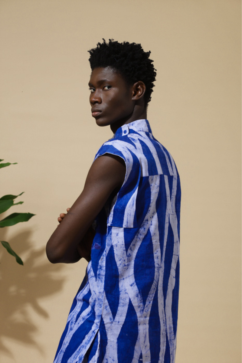 Back view of male model in Cute Saint Irin-Ajo royal blue and white bold stripe batik sleeveless shirt with arms crossed, styled with tropical plant