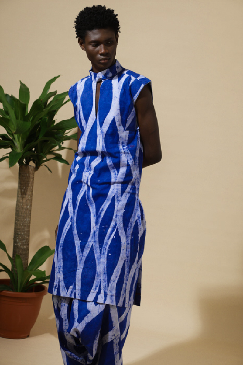 Shirtless male model wearing Cute Saint Irin-Ajo cobalt blue and white stripe batik balloon harem pants and shirt with drawstring ankles, styled with potted plant and scattered fruits